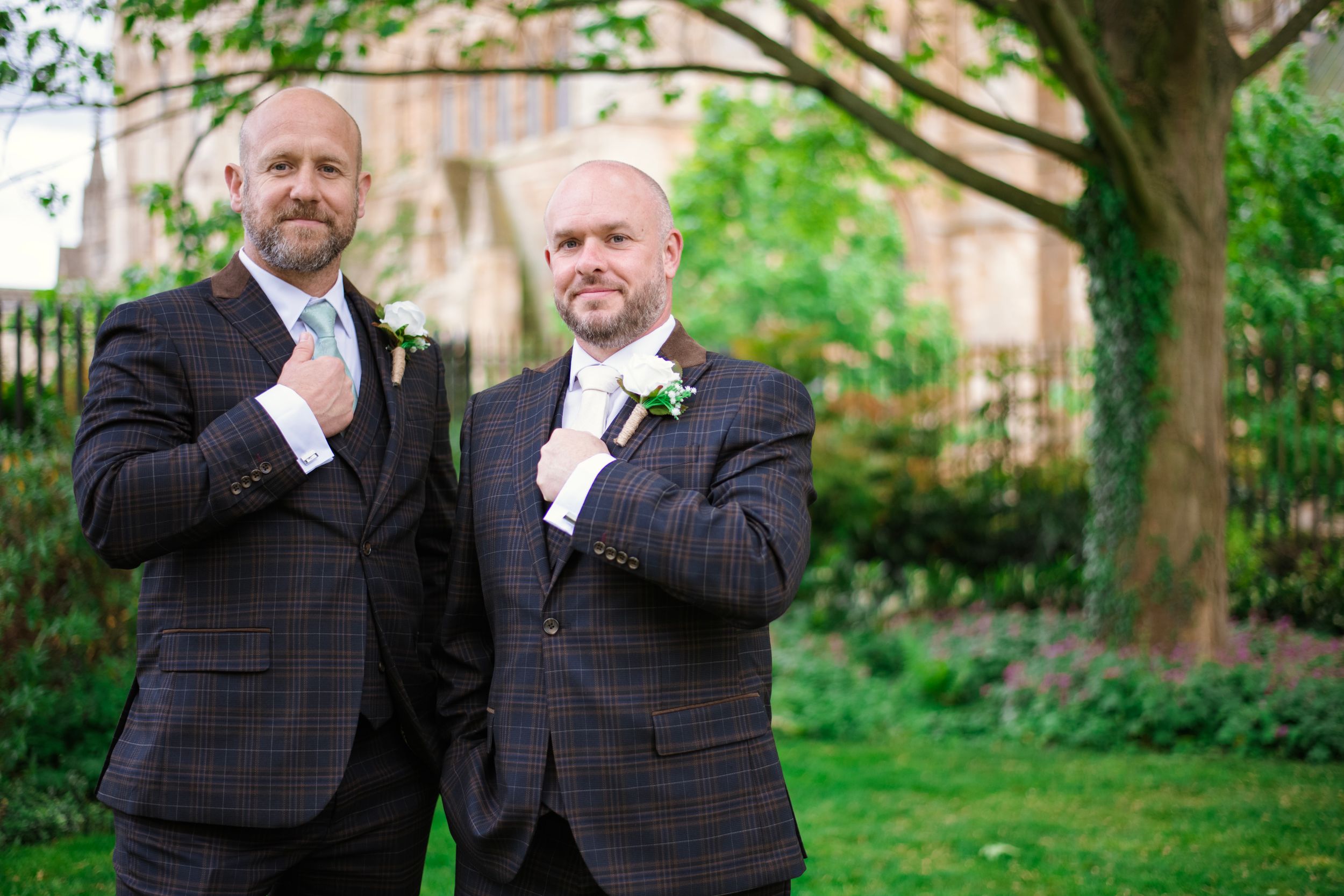 Two people in matching plaid suits and white boutonnieres pose together in a garden setting at a wedding celebration.