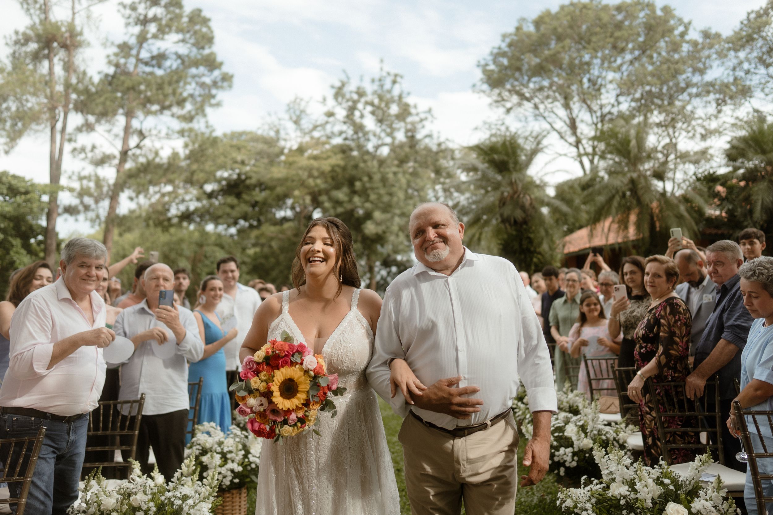 A sunny outdoor wedding ceremony with a couple walking down the aisle as guests look on under tall trees.