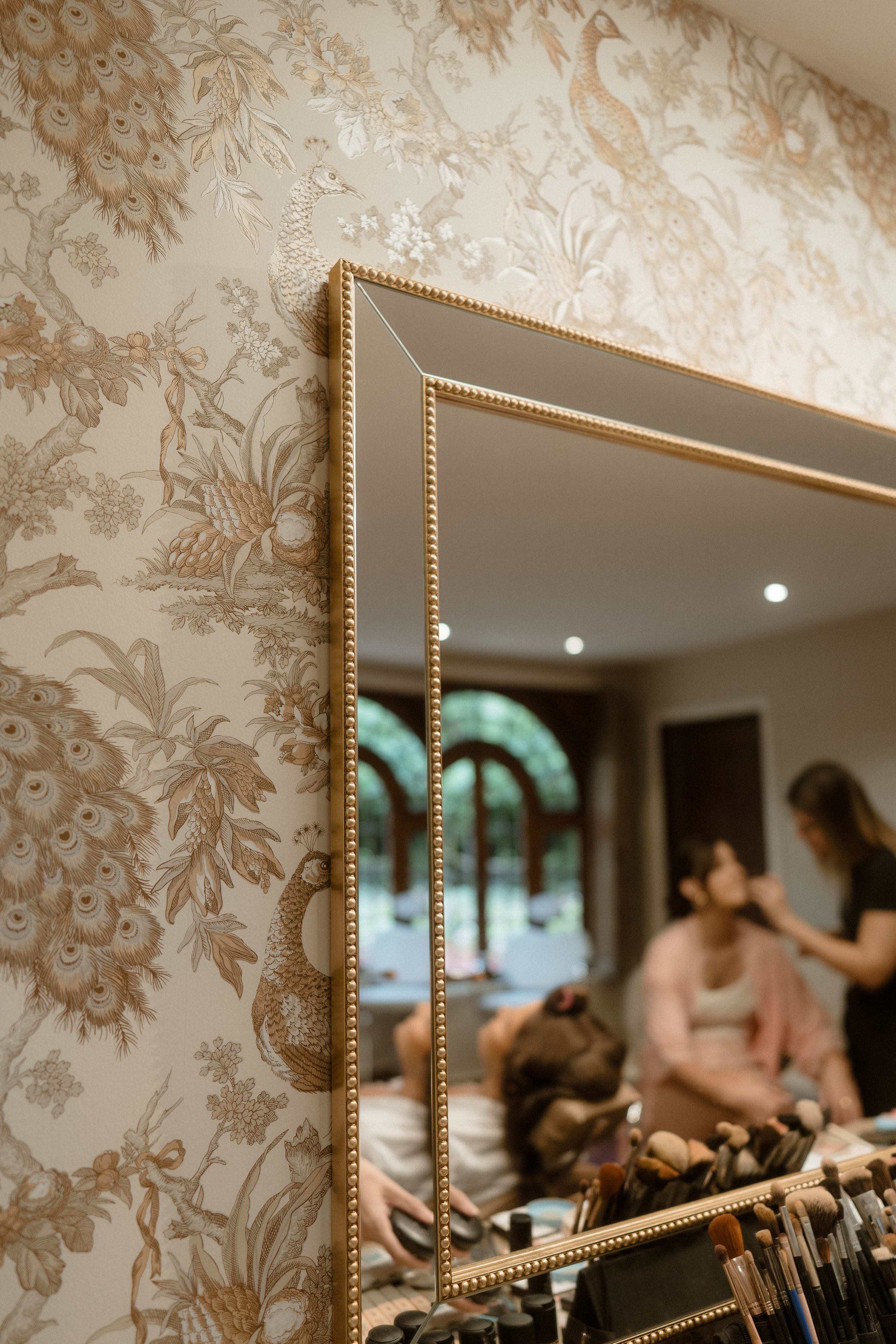 Ornate gold-framed mirror reflecting room with floral wallpaper and subtle lighting.