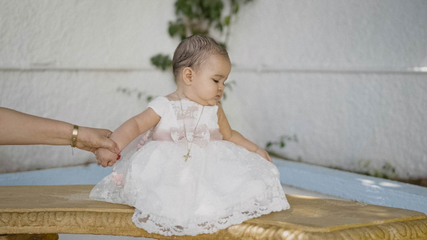 A baby in a white lace dress sits on a golden surface while being supported by someone's hand.