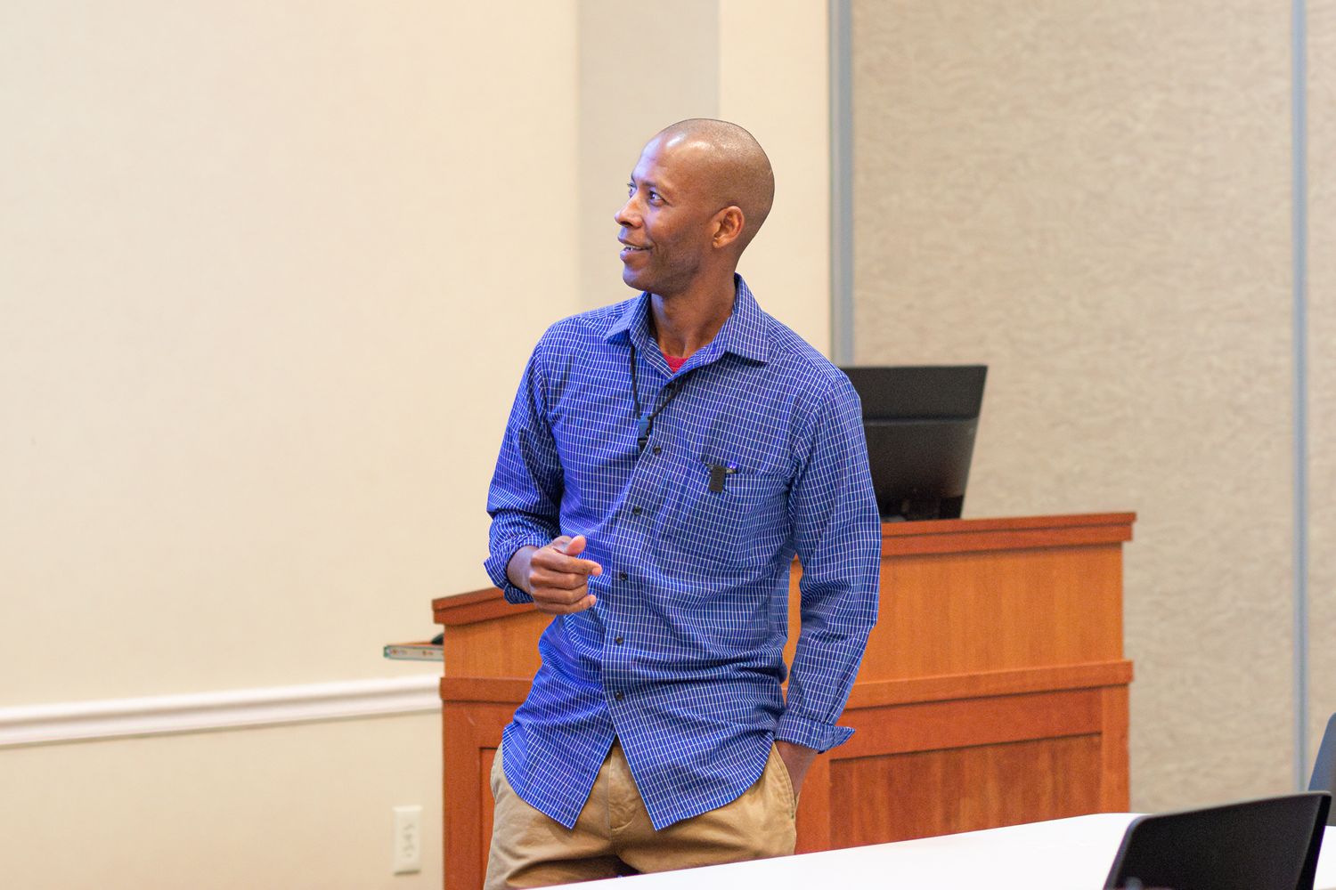 Speaker in blue button-down shirt smiling while giving presentation at podium in conference room setting.