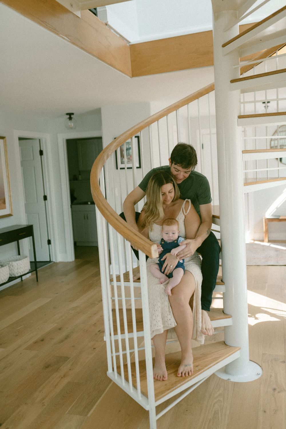 A couple sits together on white spiral stairs inside a bright modern home with wood flooring and natural light.