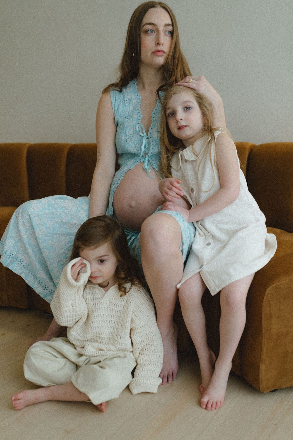 A pregnant figure in turquoise dress sits on a brown couch with two small children in neutral-colored outfits.