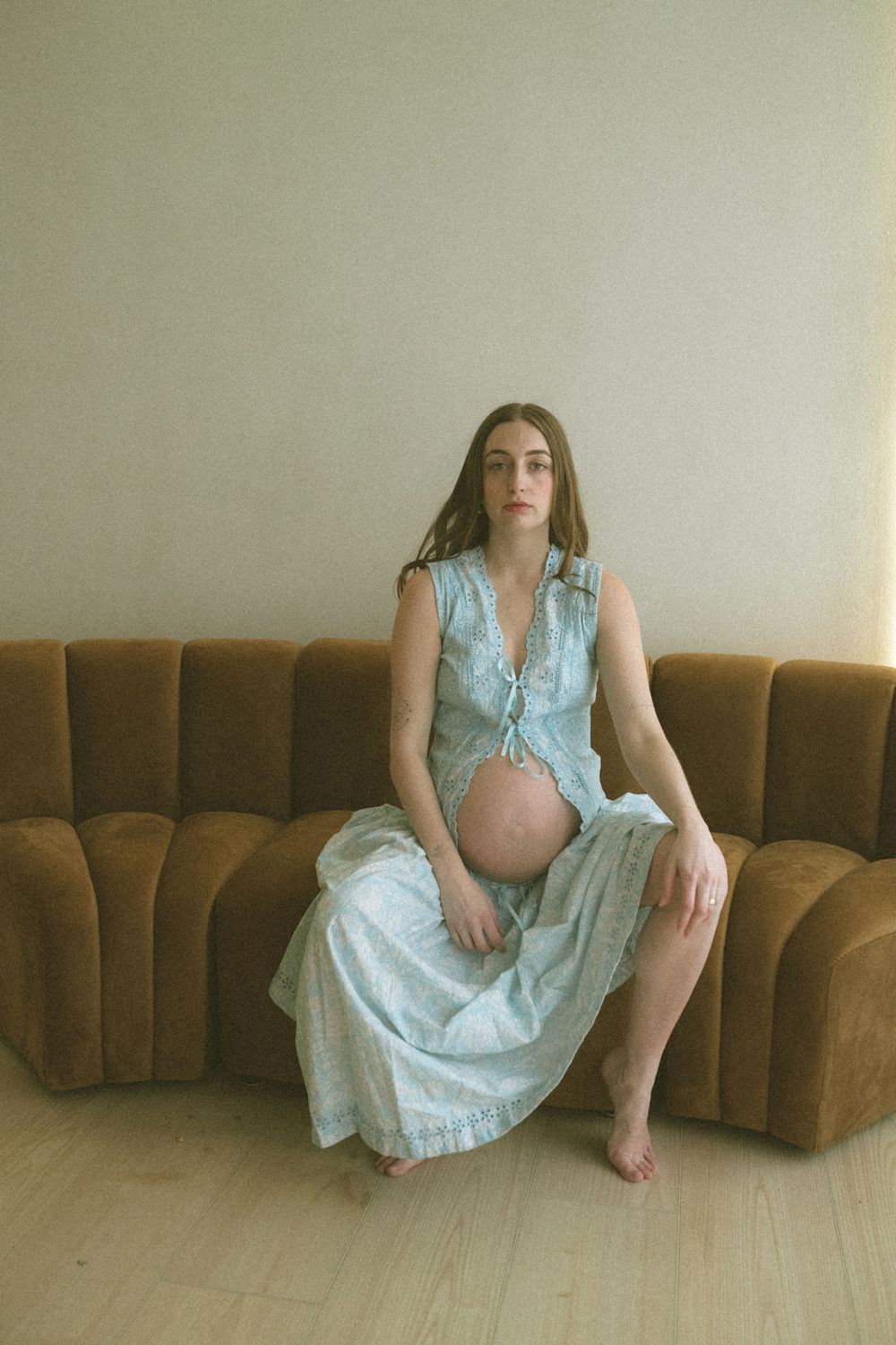 A person in a denim vest and long skirt sits on a brown velvet couch in a minimalist interior setting.