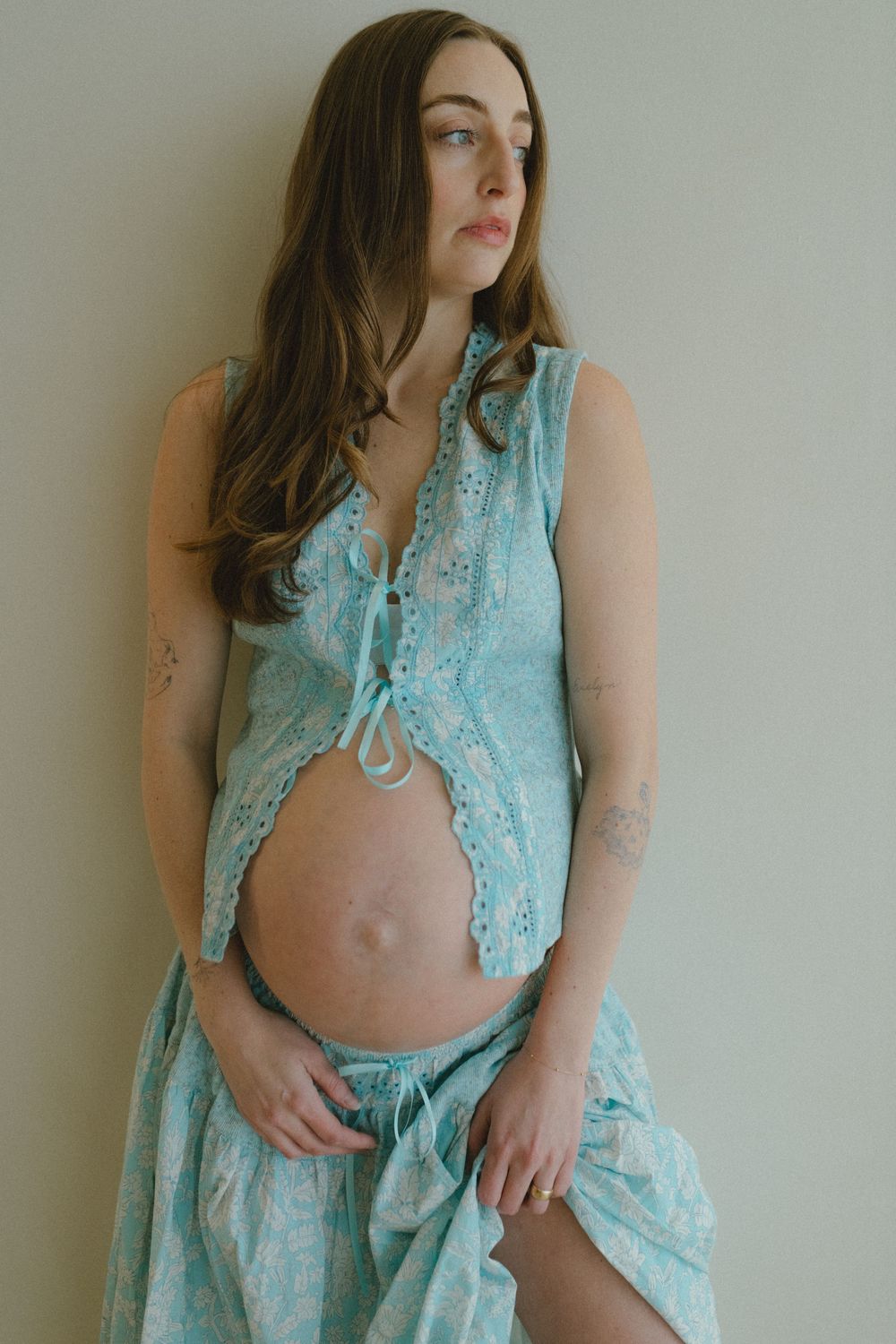 A pregnant woman in a flowing turquoise lace dress poses against a white wall during a maternity photoshoot.