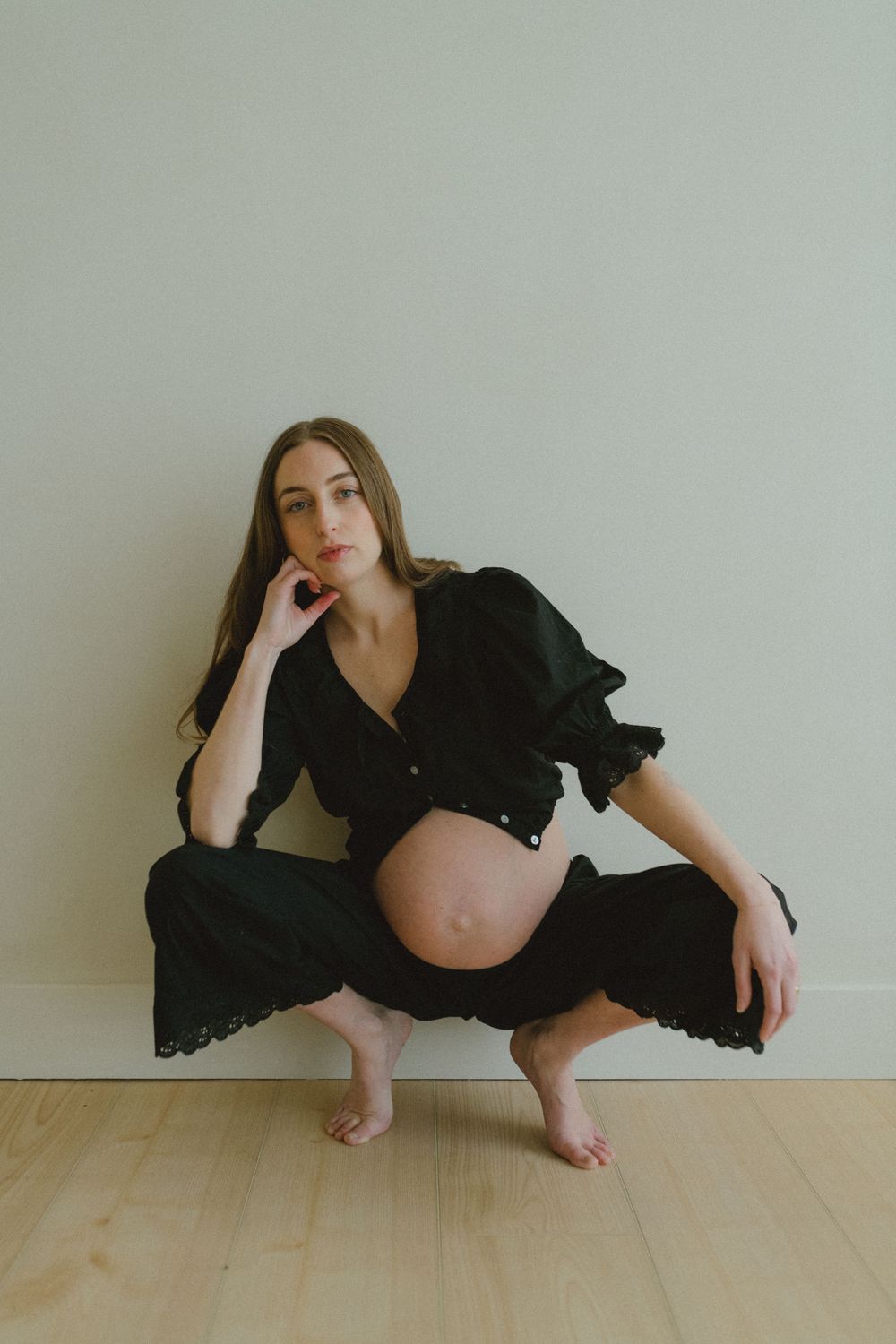 Maternity portrait featuring a relaxed pose in a flowing black dress against a minimalist white wall and wood floor.