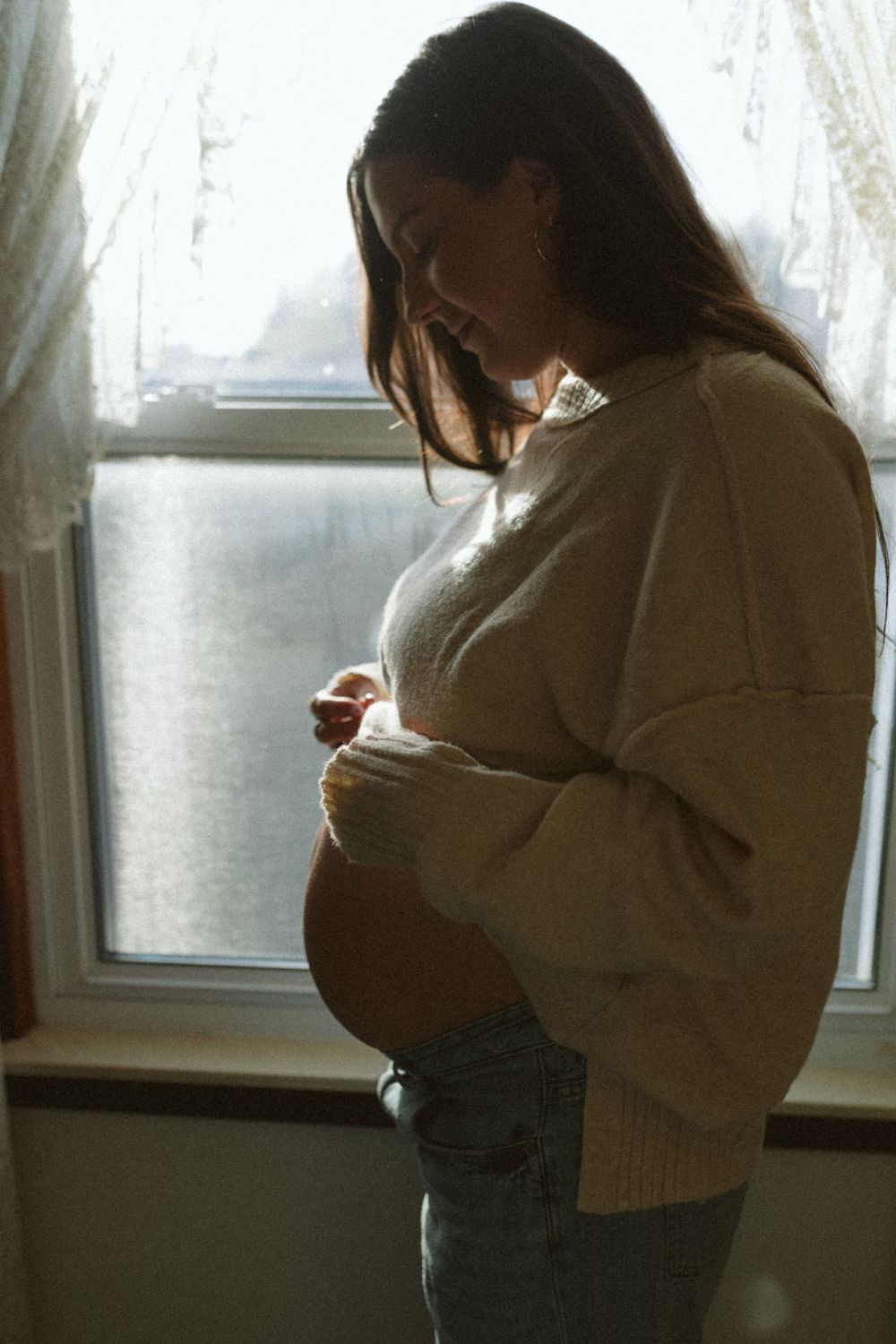 A person in a cream sweater and dark pants stands by a window in a dimly lit room looking down at something.