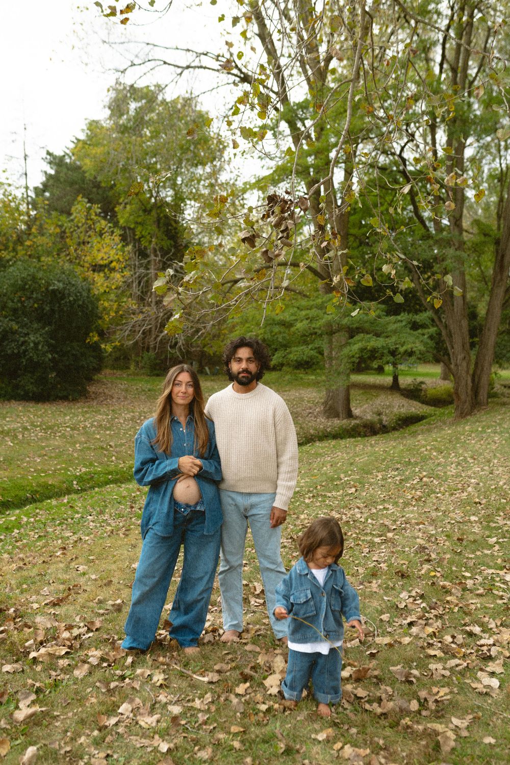 A family wearing denim outfits poses together in a park with autumn leaves scattered on the ground.