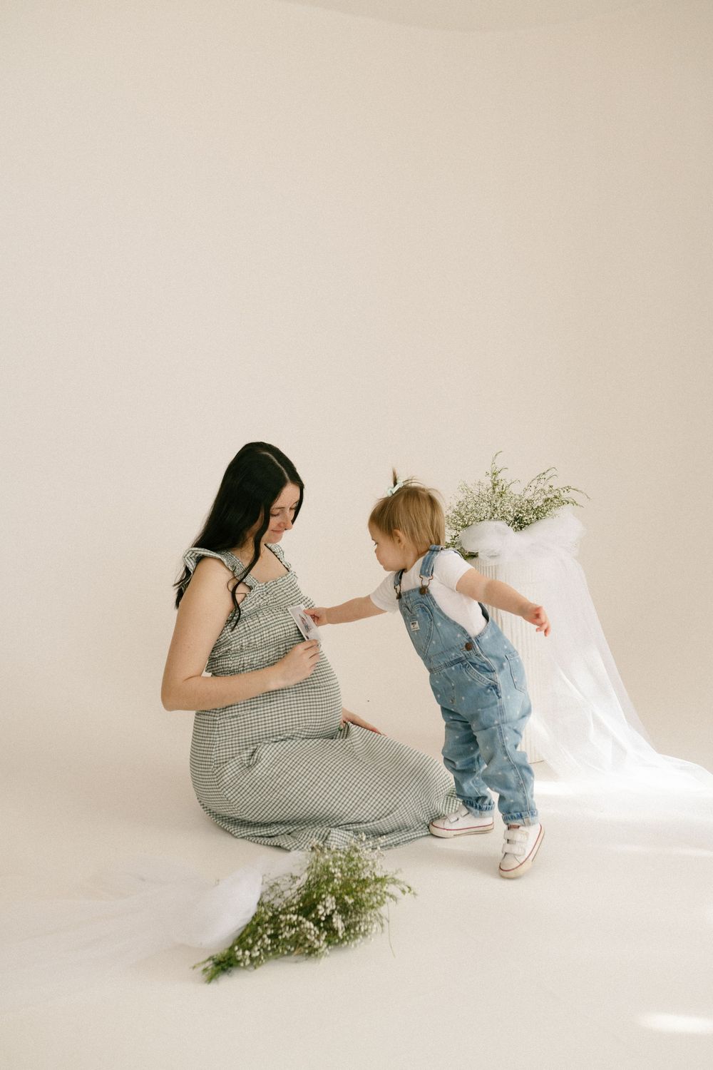 A maternity session in a minimalist studio with baby breath flowers and white draping as a toddler touches a pregnant woman.