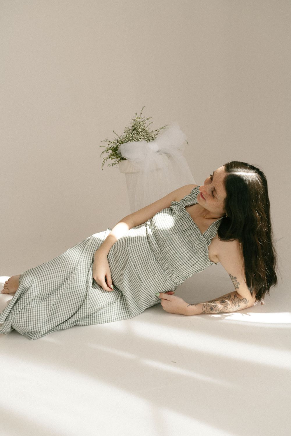 Woman in checkered dress poses elegantly on floor with sunlight and potted plant in minimalist setting.