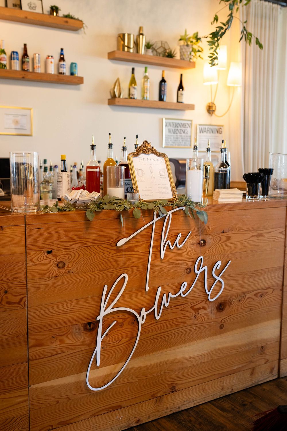 Rustic wooden bar with floating shelves displaying bottles and white neon 'The Booze' sign decorated with greenery.