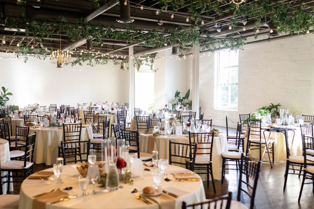 Wedding reception venue with ivory brick walls, greenery decor, round tables with gold chiavari chairs and table settings.