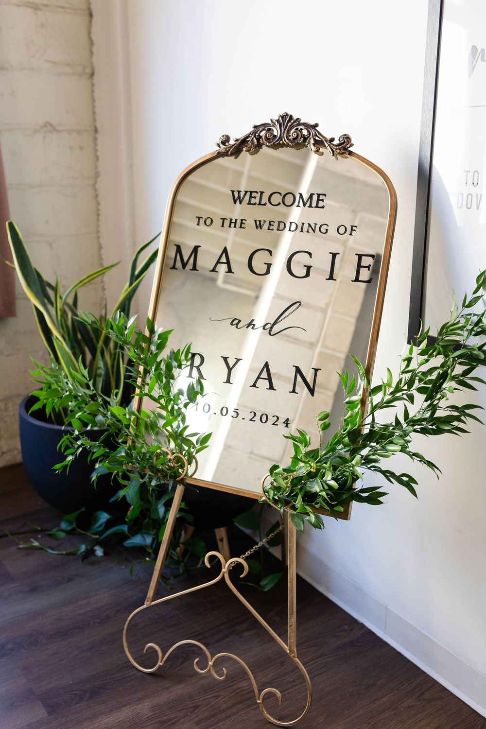 An elegant wedding welcome sign decorated with greenery displays 'Welcome to the Wedding of Maggie and Ryan' on a gold stand.