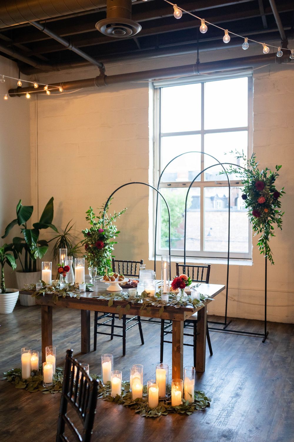 Romantic wedding reception setup with rustic wooden table, candles, and floral arches in an industrial-style venue.