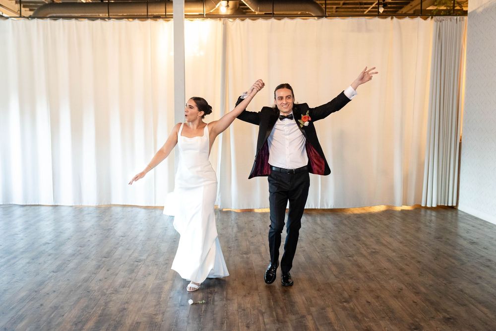 A newlywed couple joyfully dances together in an elegant venue with white curtains and wooden floors.