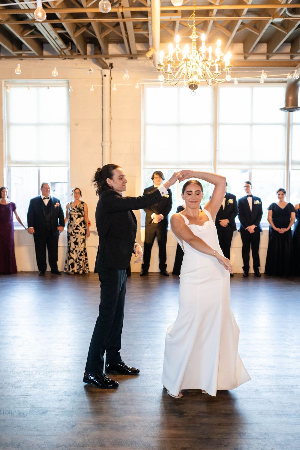 A couple performs their first dance in an elegant venue with wooden floors and a crystal chandelier overhead.