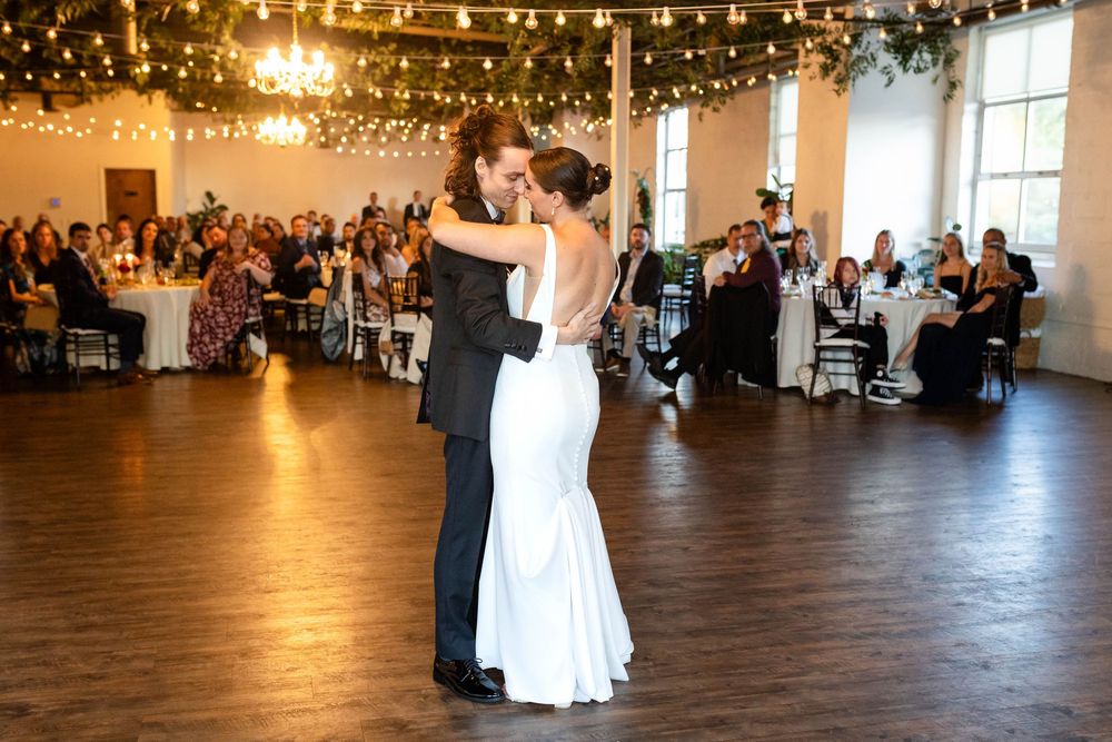 A couple shares their first dance on a wooden floor under string lights at a wedding reception with guests watching.