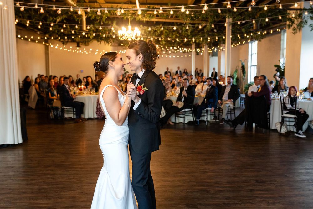 A couple shares their first dance at a wedding reception under string lights and a chandelier in an elegant ballroom.