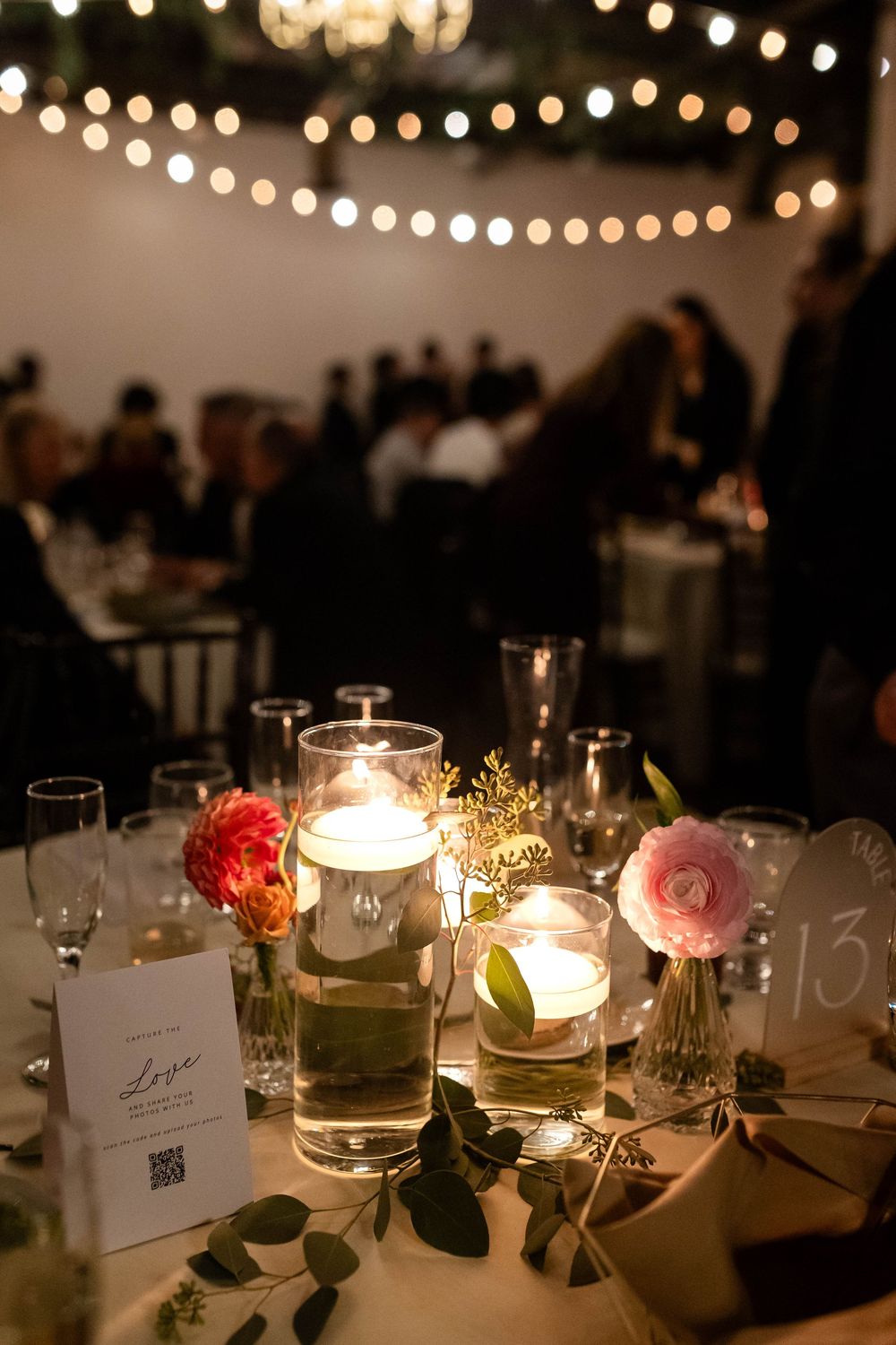 Elegant wedding reception table with glowing candles, string lights overhead, and delicate floral centerpieces in soft lighting.