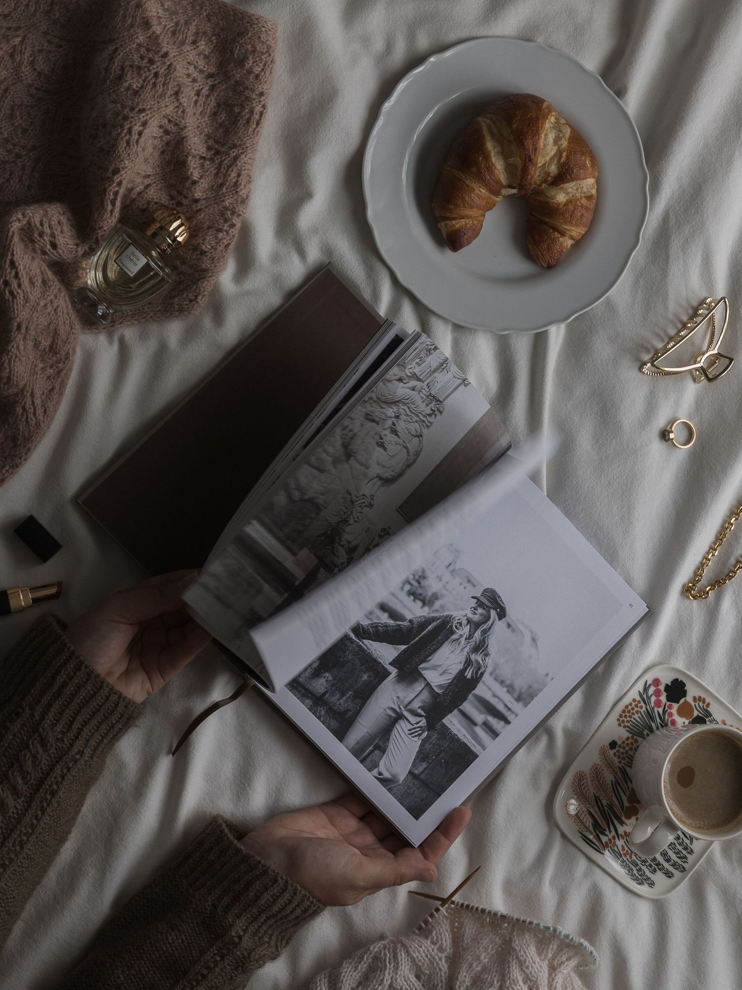 Viihtyisä flatlay, jossa on vintage-valokuvia, croissant, kahvikuppi ja koruja valkoisella vuodevaatteella.