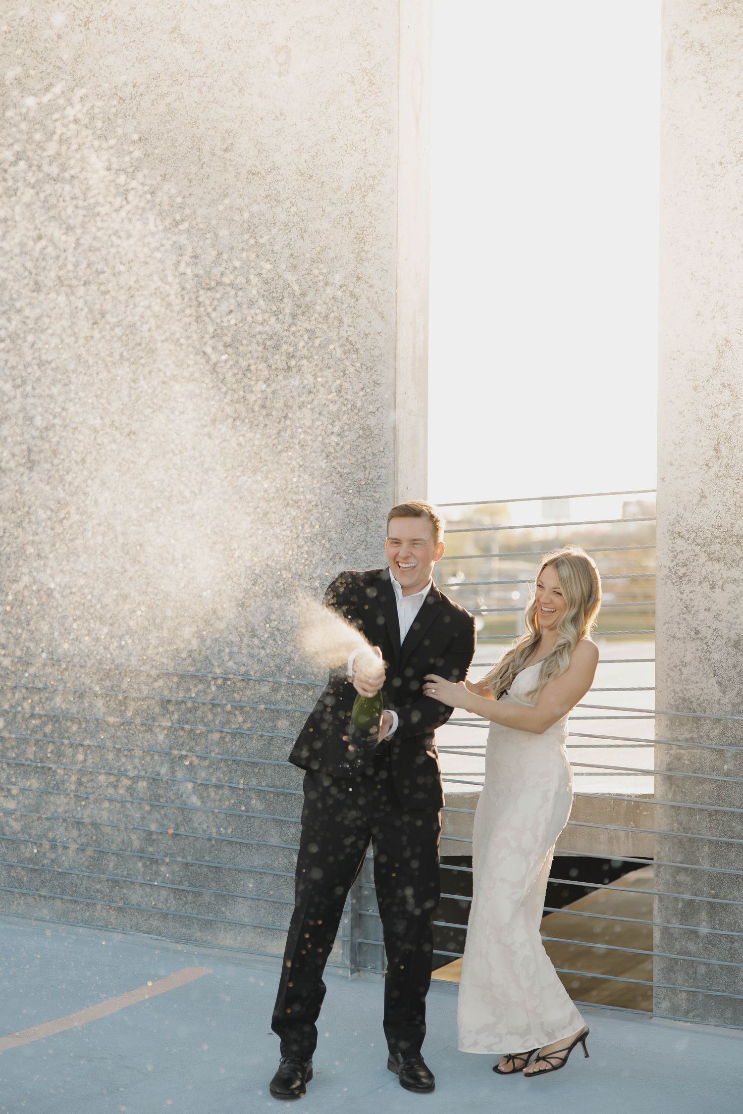 A couple in formal attire celebrates with champagne spray against a light wall backdrop.