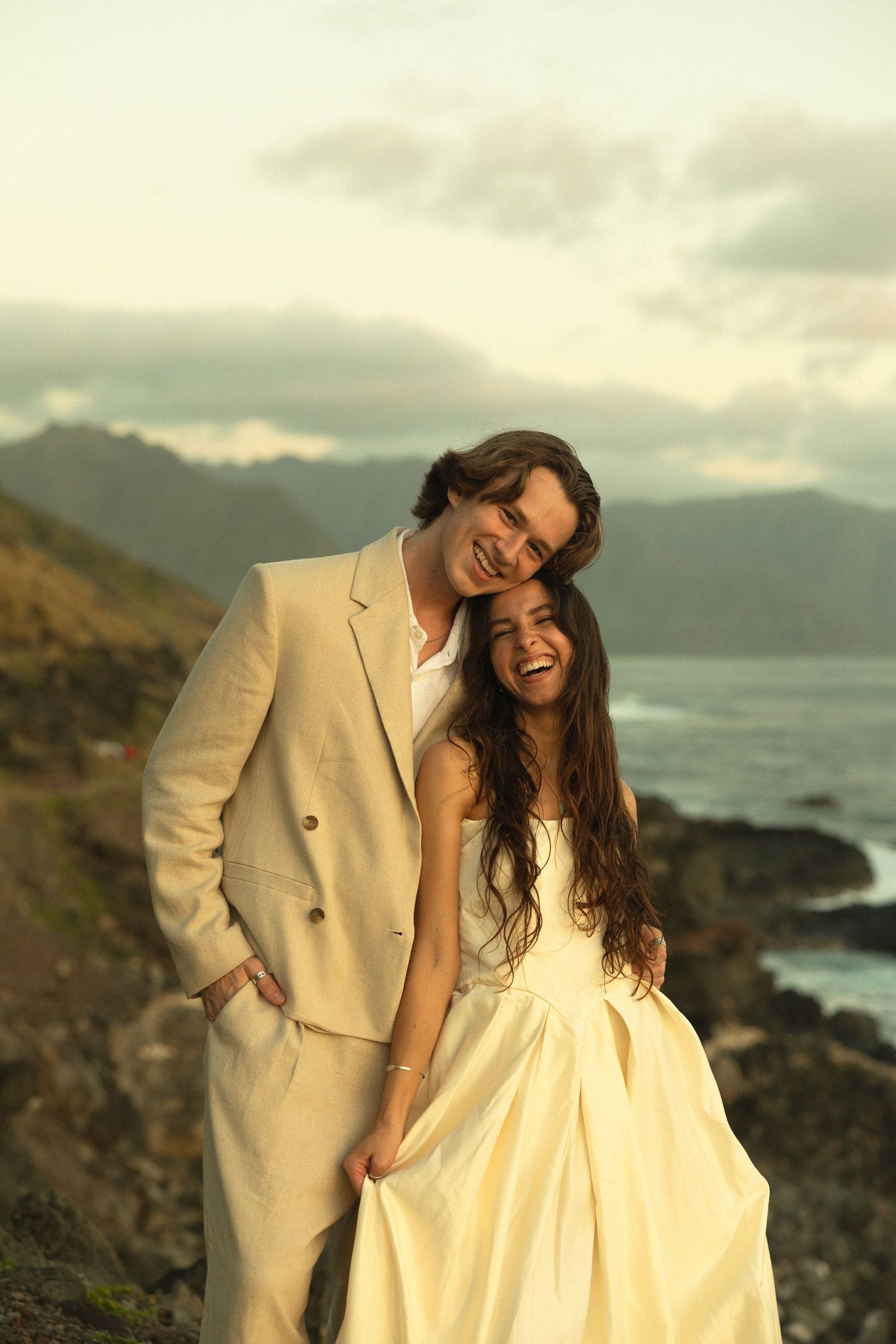 A romantic couple in formal attire embraces on a rocky coastline at sunset with mountains and ocean in the background.