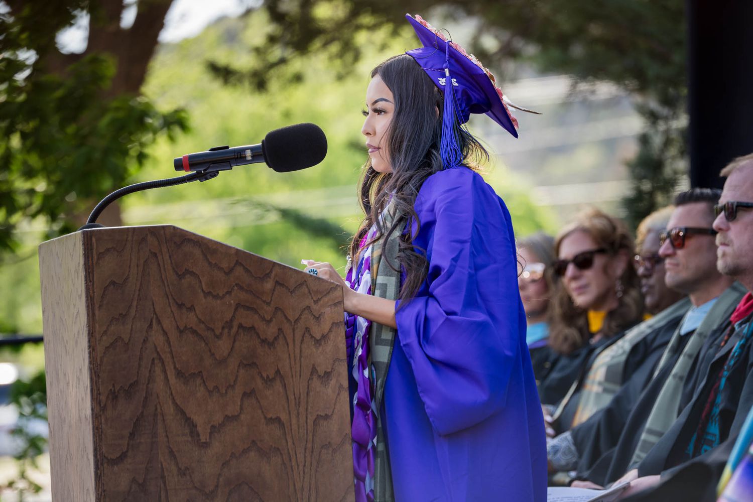 A graduate in a purple cap and gown delivers a speech at a podium during an outdoor commencement ceremony.