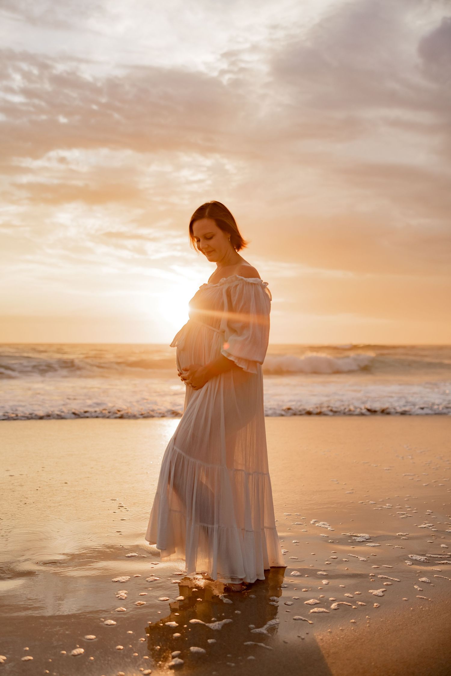 Family wearing white and beige clothing poses together on beach at sunset while young child dances.