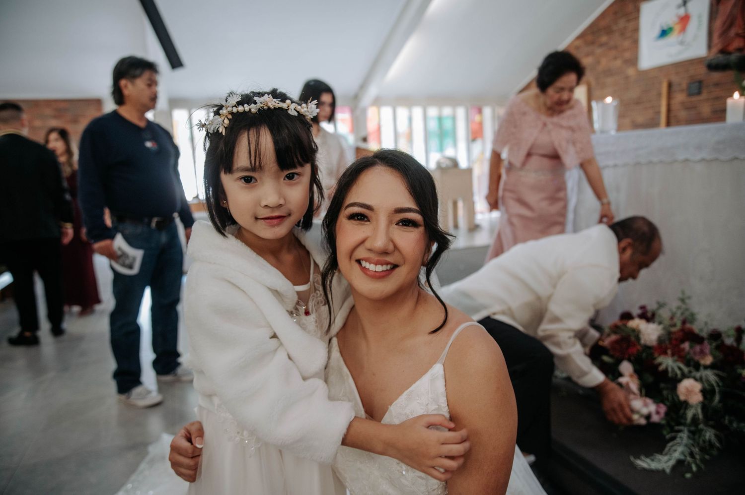 A bright and joyful celebration at an indoor wedding venue with white decor and floral arrangements in the background.