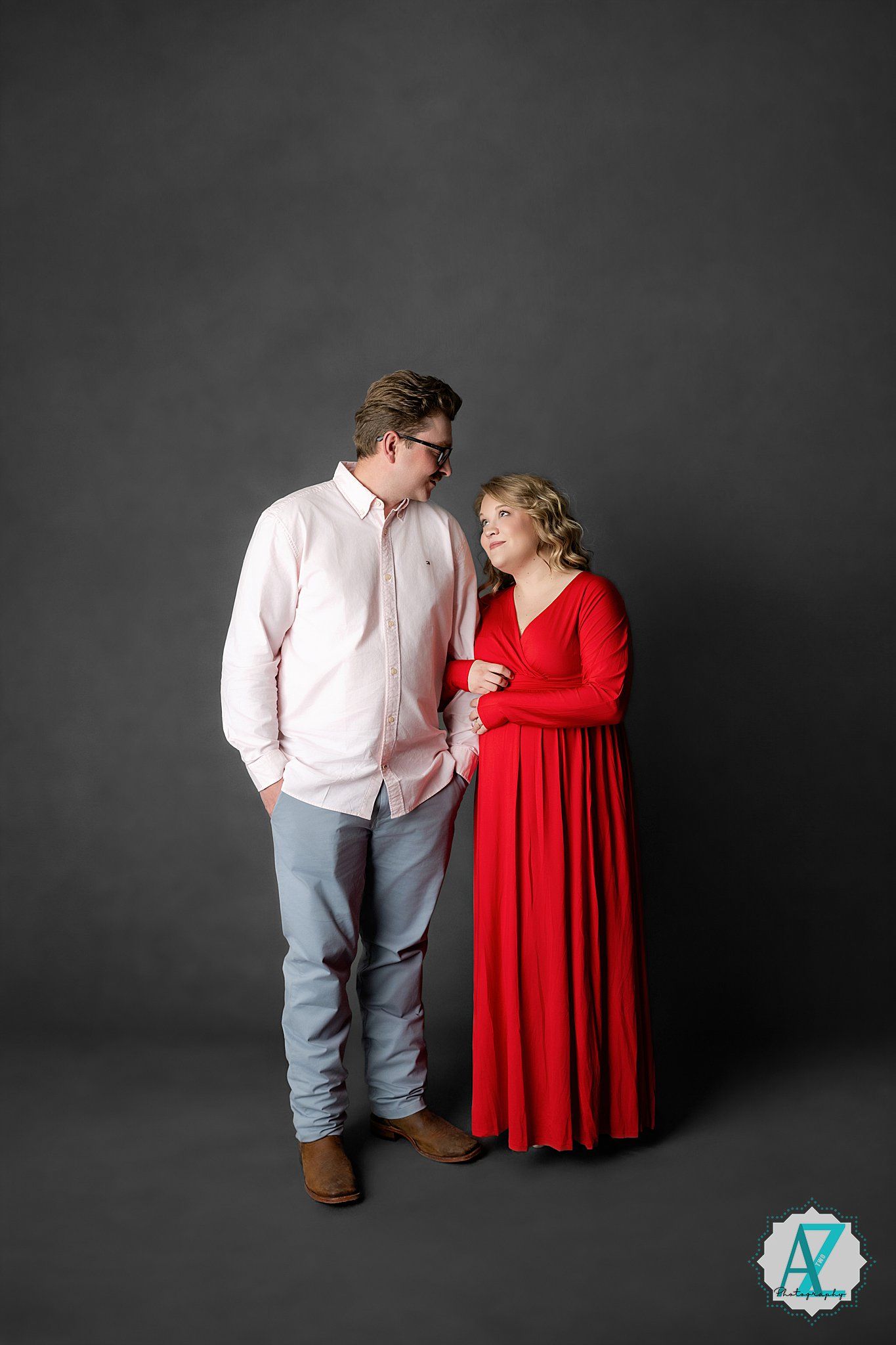A couple poses together against a dark backdrop, with the woman wearing a flowing red dress and the man in casual attire.
