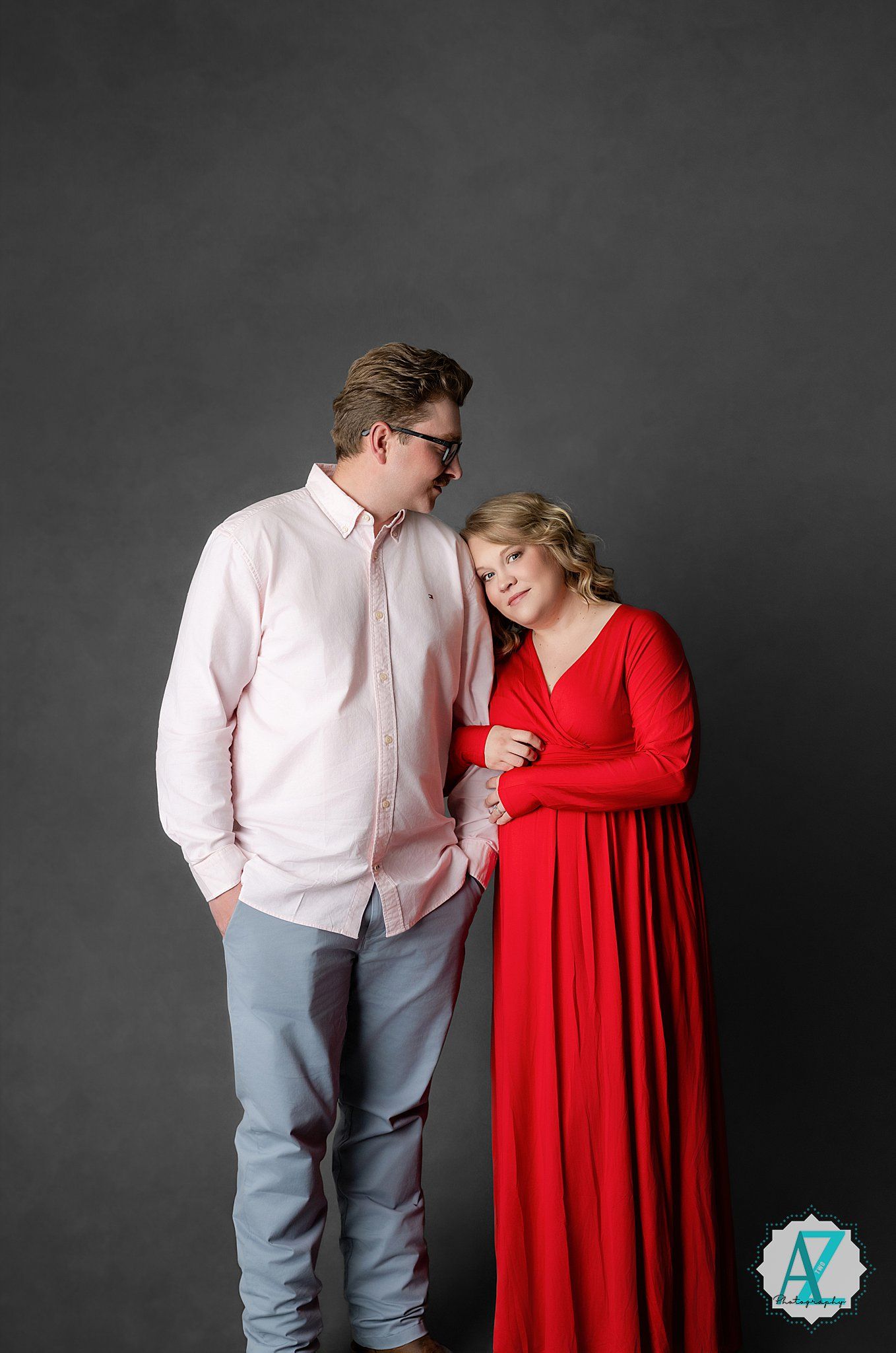 A couple poses together against a gray background, with one person in a white shirt and jeans and another in a flowing red dress.