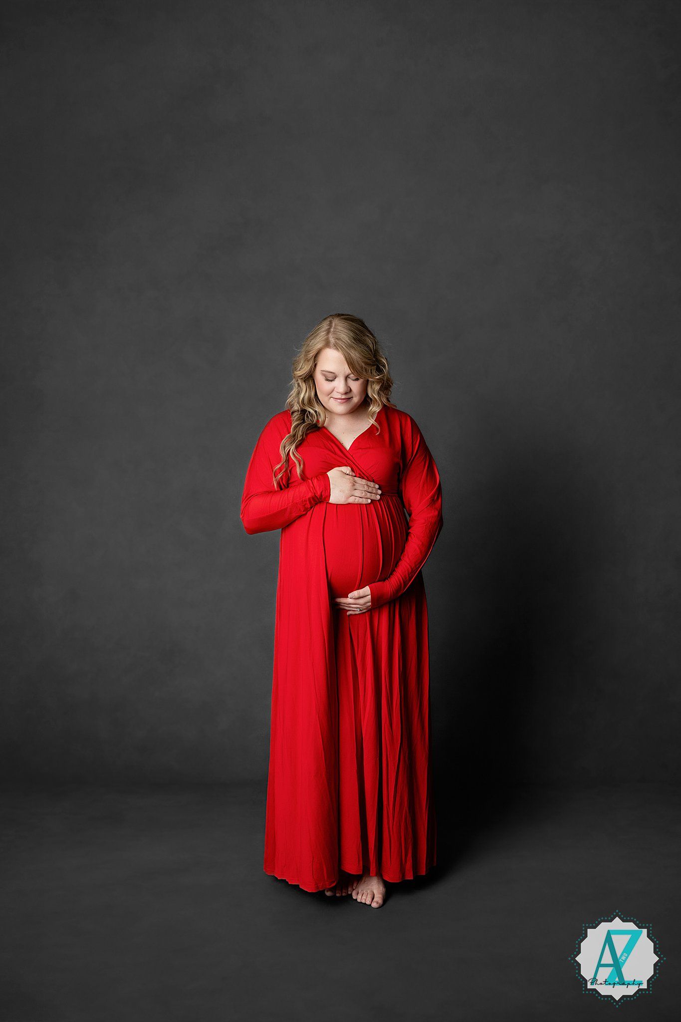A person in a flowing red long-sleeved maxi dress poses against a dark grey background in a maternity photo.
