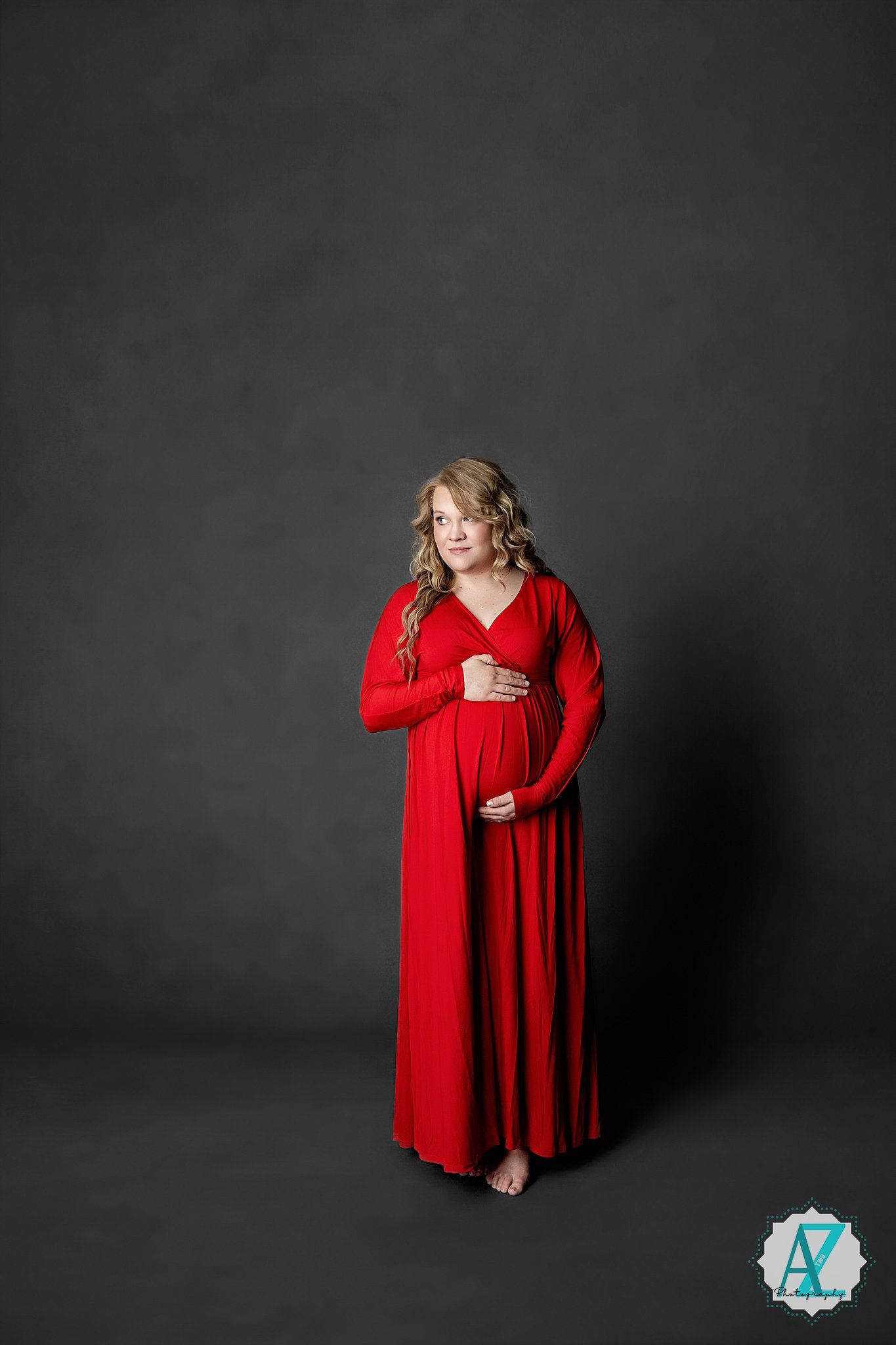 A figure in a flowing red long-sleeved maxi dress stands against a dark grey backdrop in an elegant maternity pose.