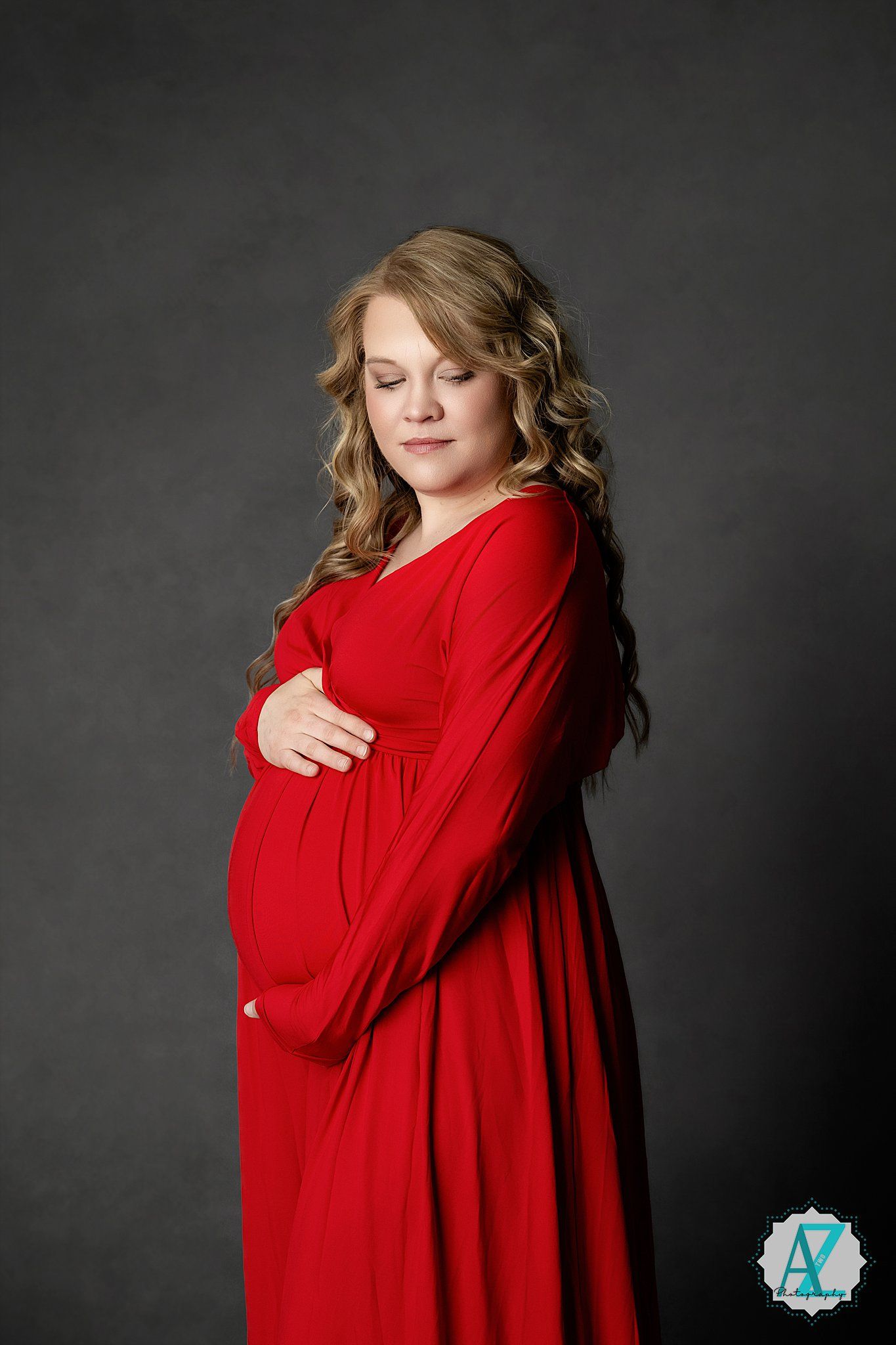A pregnant woman in a long red dress poses sideways against a dark background while touching her baby bump.