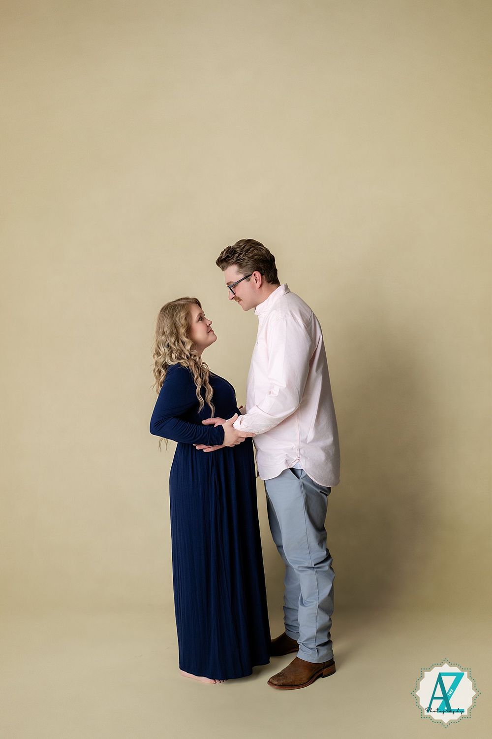 A couple embraces in a studio photo, with her wearing a navy maxi dress and him in a white shirt and jeans.