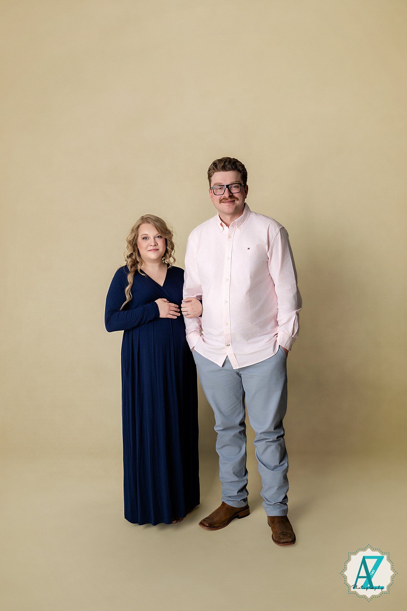 A couple poses together against a beige backdrop, with a woman in a navy dress and a man in a white shirt and khakis.