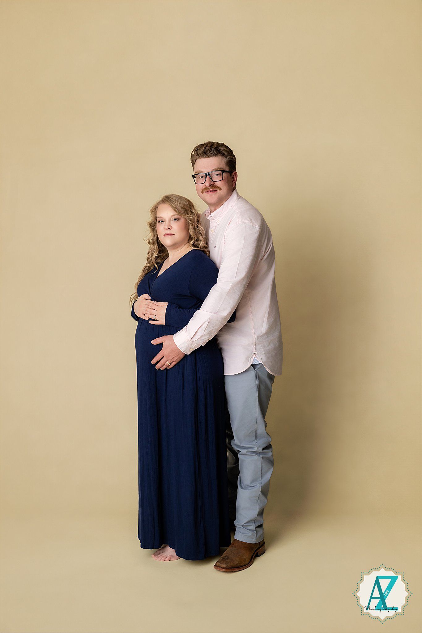 A couple poses together against a beige backdrop, with the woman wearing a navy blue maxi dress and the man in a white shirt.