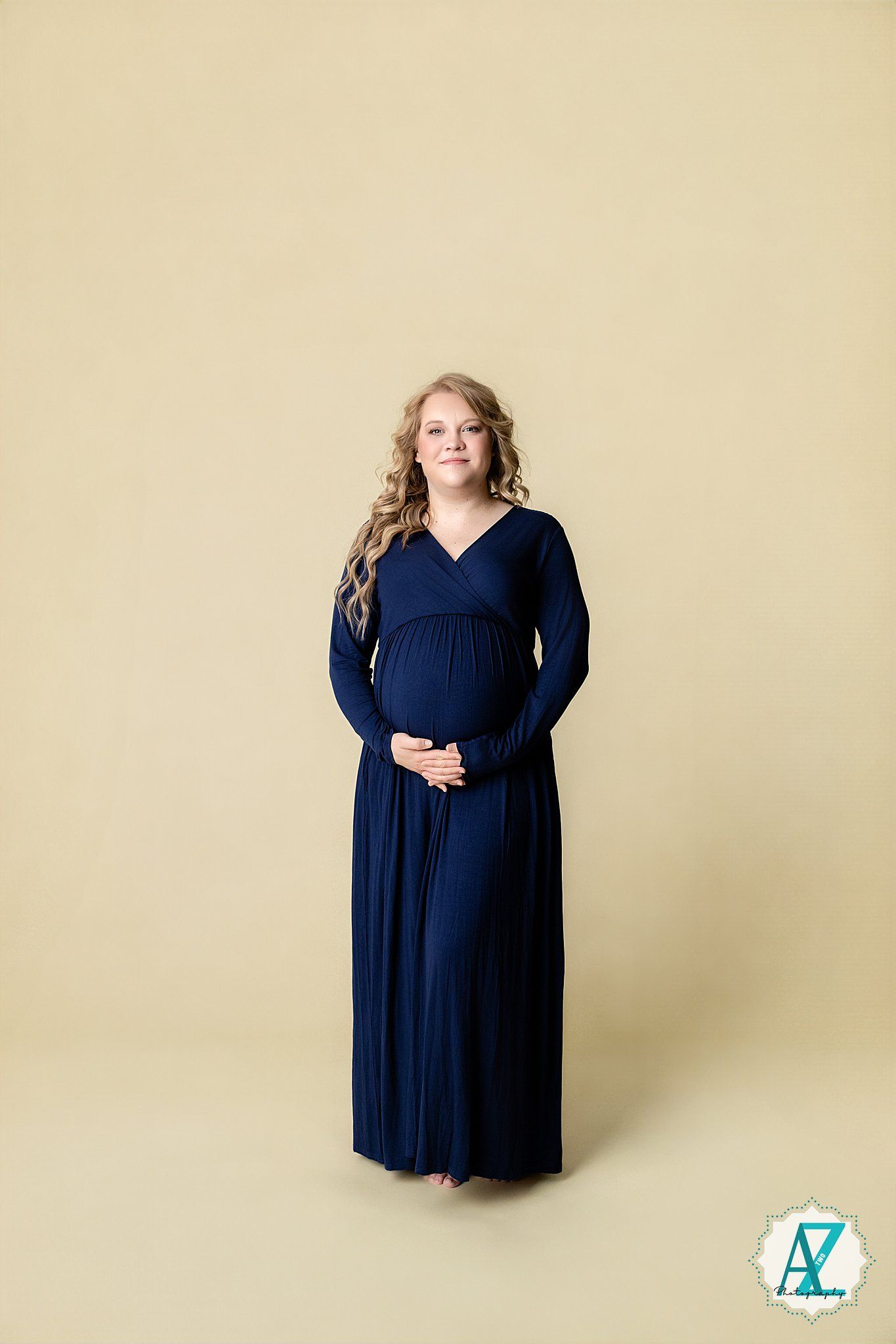 Expectant mother wearing a long navy blue maxi dress with v-neck and long sleeves against beige background.