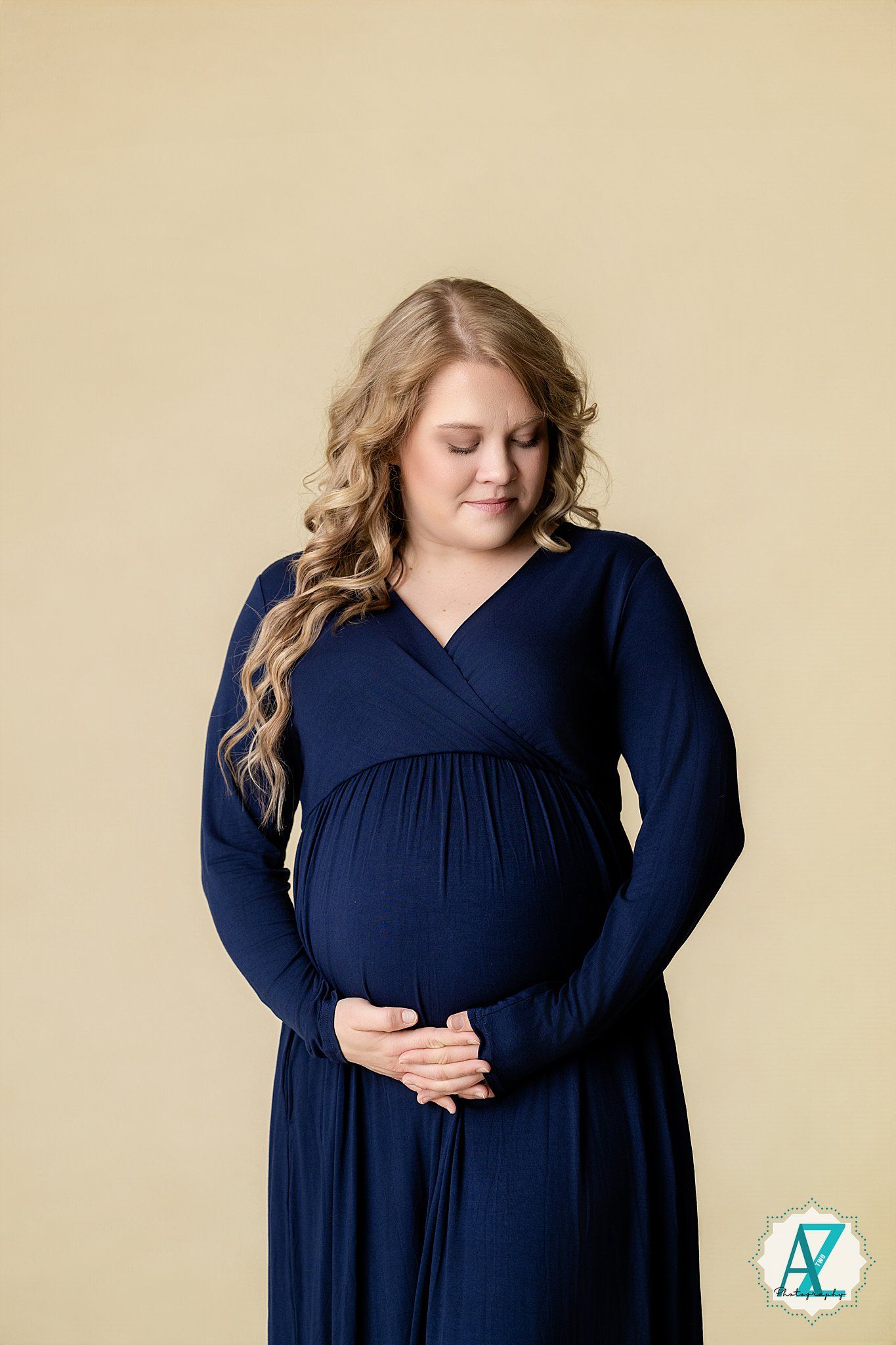 A pregnant woman in a long navy dress smiles while cradling her baby bump against a beige background.