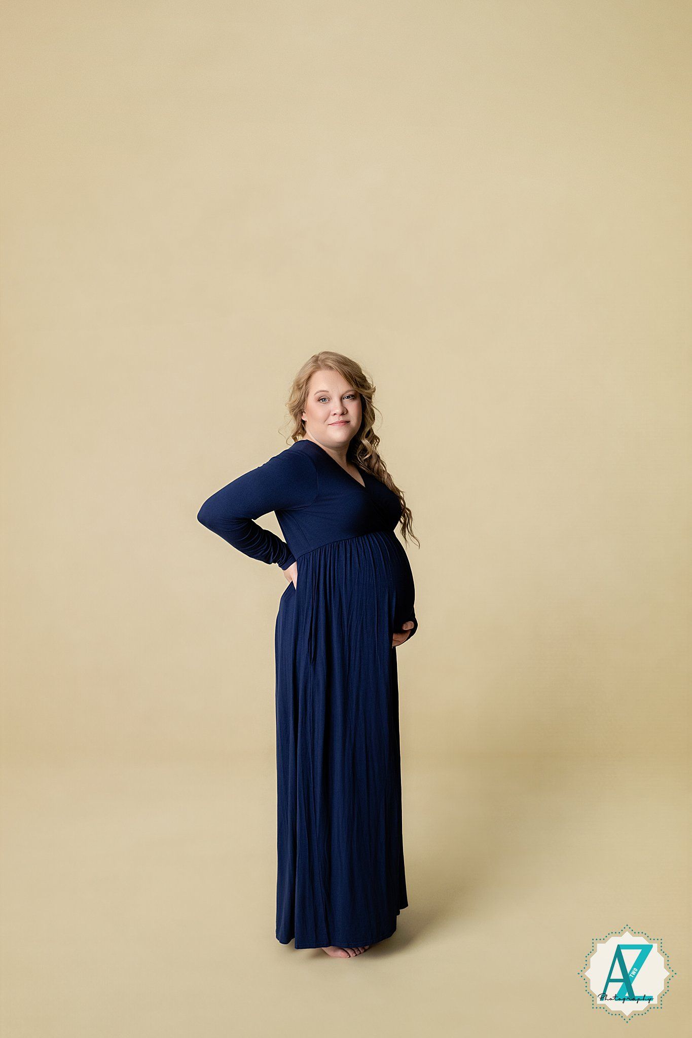A pregnant woman in a flowing navy maxi dress poses with hand on hip against a neutral backdrop.