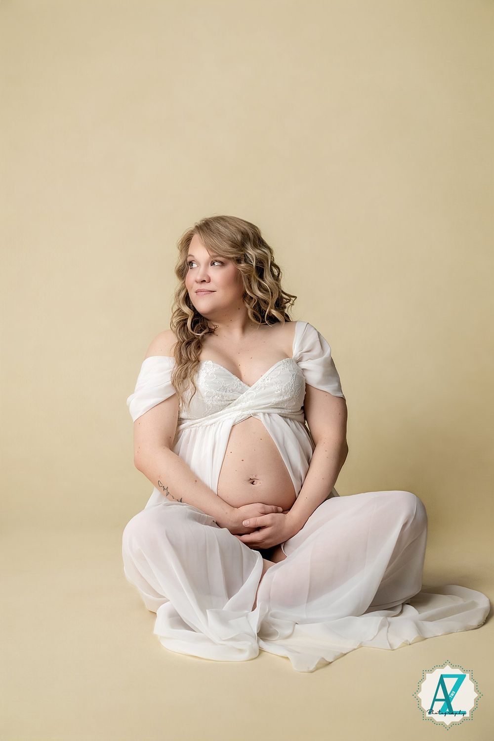 A maternity photoshoot featuring a person in a flowing white dress sitting on the floor against a beige background.