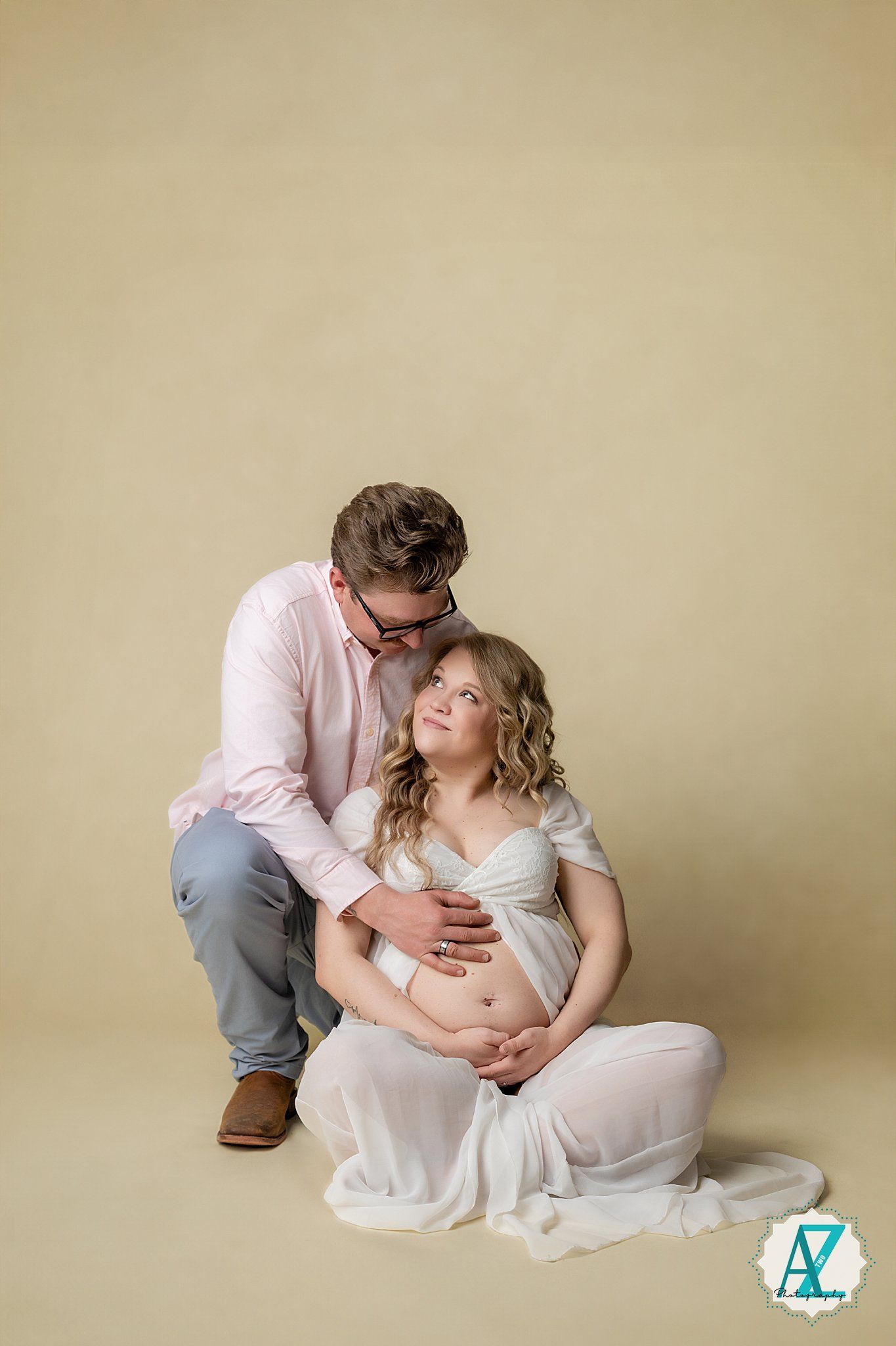 A tender maternity photoshoot featuring a couple in white and pink against a beige backdrop with soft lighting.