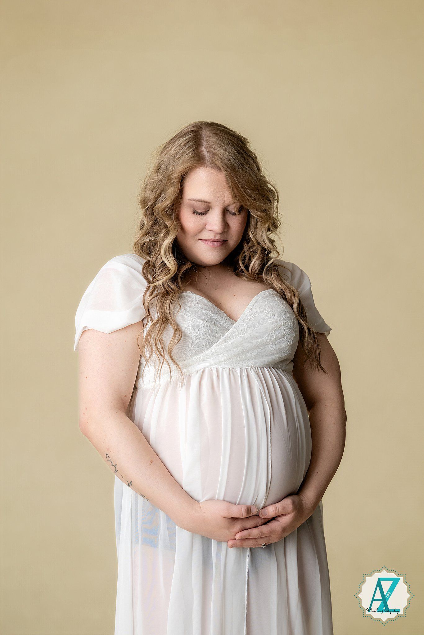 Expectant mother in flowing white maternity dress smiles peacefully with hands cradling her baby bump against beige backdrop.