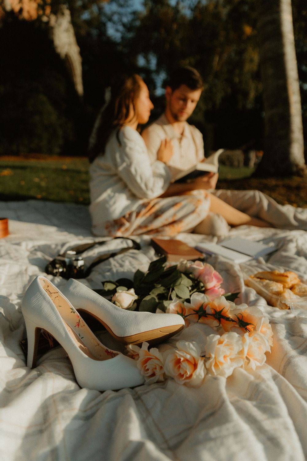 White bridal shoes and pink flowers arranged on a light blanket during an outdoor photo session.
