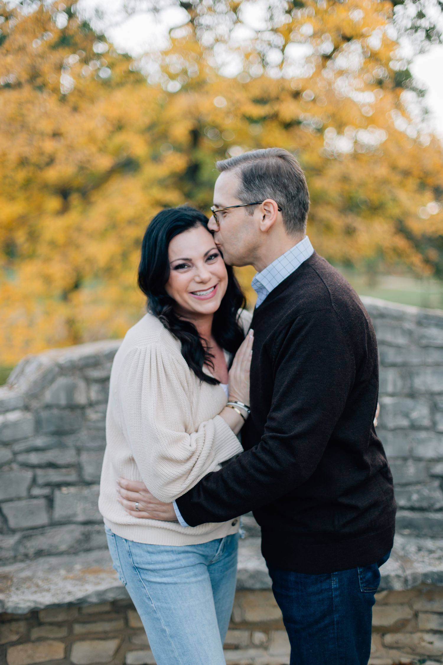 A romantic couple embraces against a stone wall with autumn foliage in the background on a sunny afternoon.