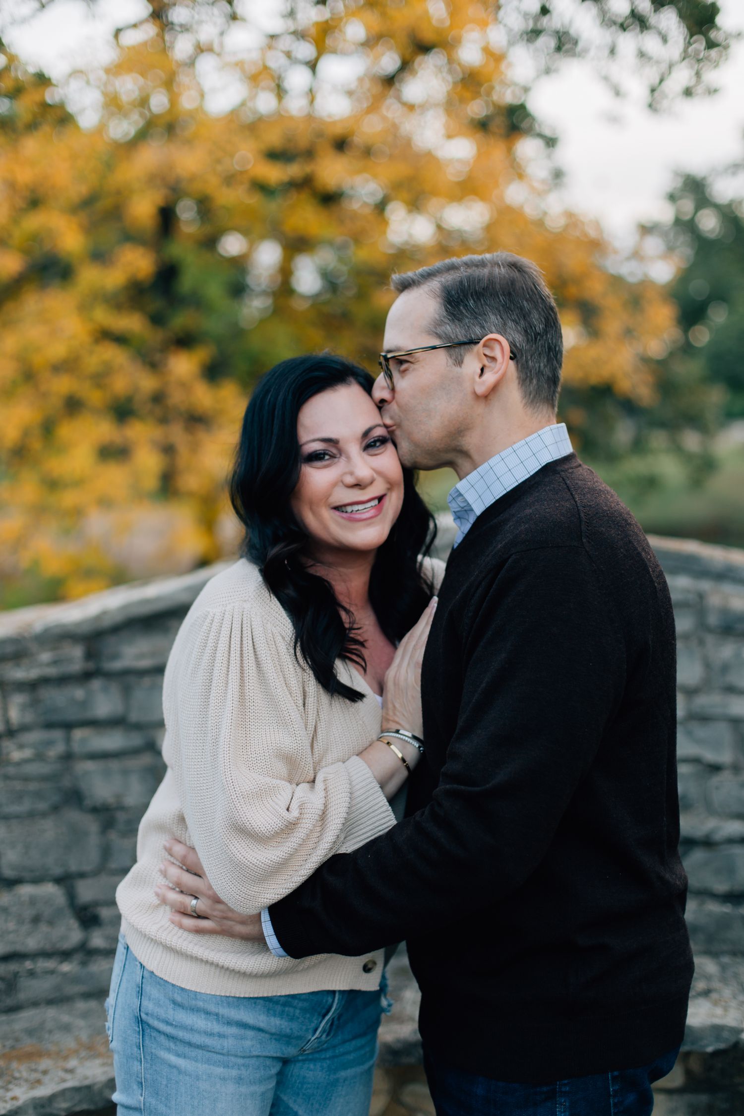 A couple share an intimate moment outdoors against a stone wall with autumn foliage in the background.