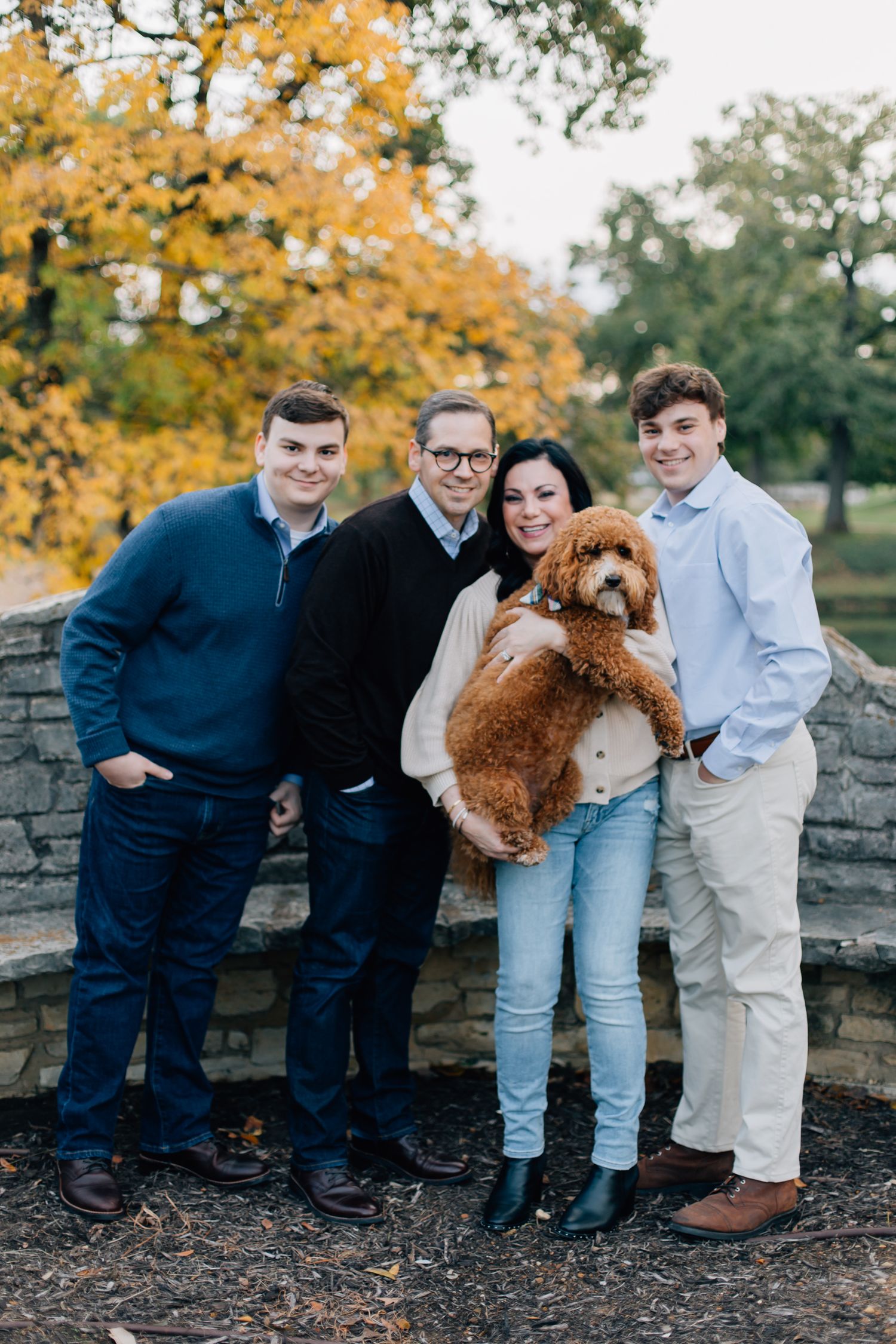 A family poses with their goldendoodle dog against a stone wall with autumn foliage in the background.