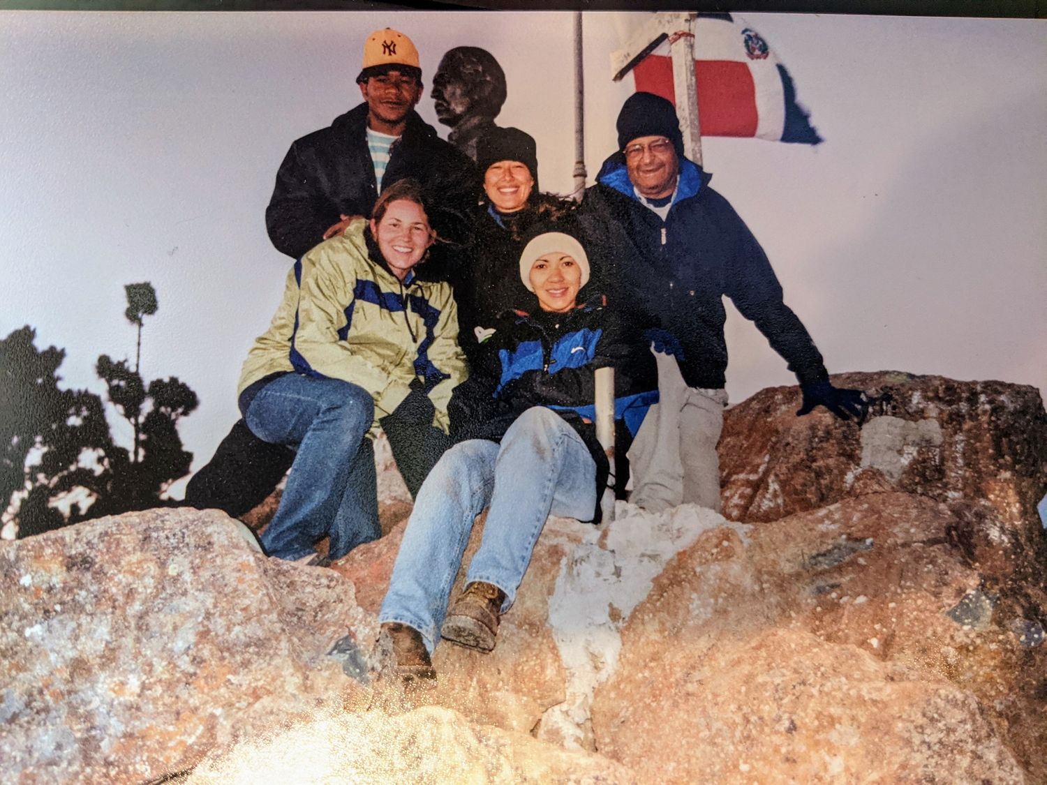 A group of people in winter clothing sit together on a large rock formation against a white background.