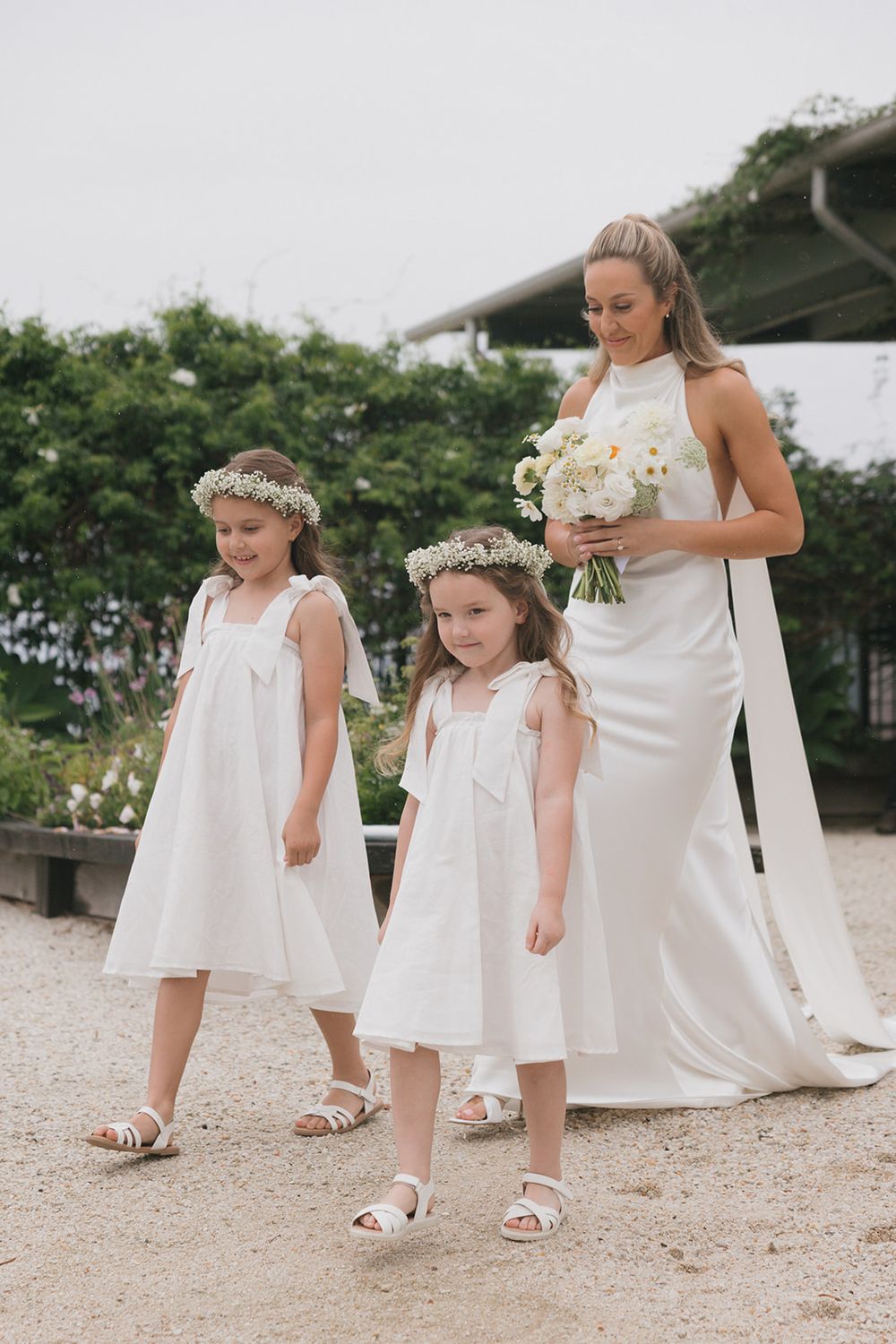 Bride in elegant white gown walks with two flower girls in matching dresses on gravel path.