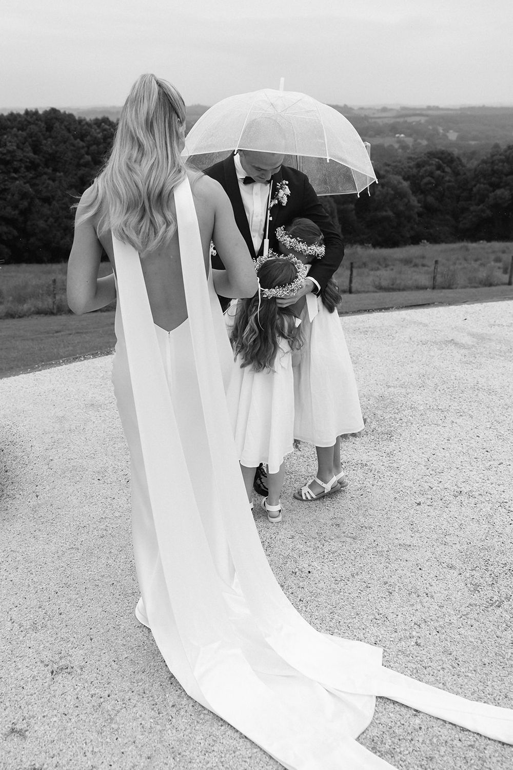 Wedding party members share intimate moment under clear umbrella during outdoor celebration.